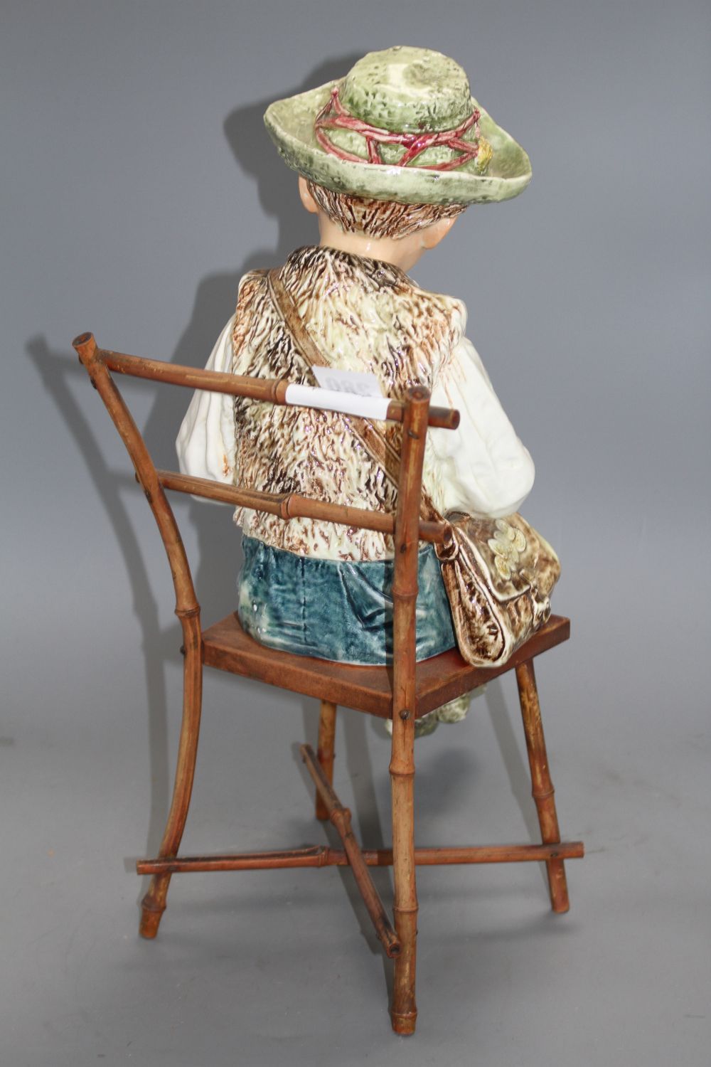 A late 19th century Austrian tin glaze pottery figure of a Tyrolean boy playing a flute, seated upon a bamboo wood chair, height 43cm